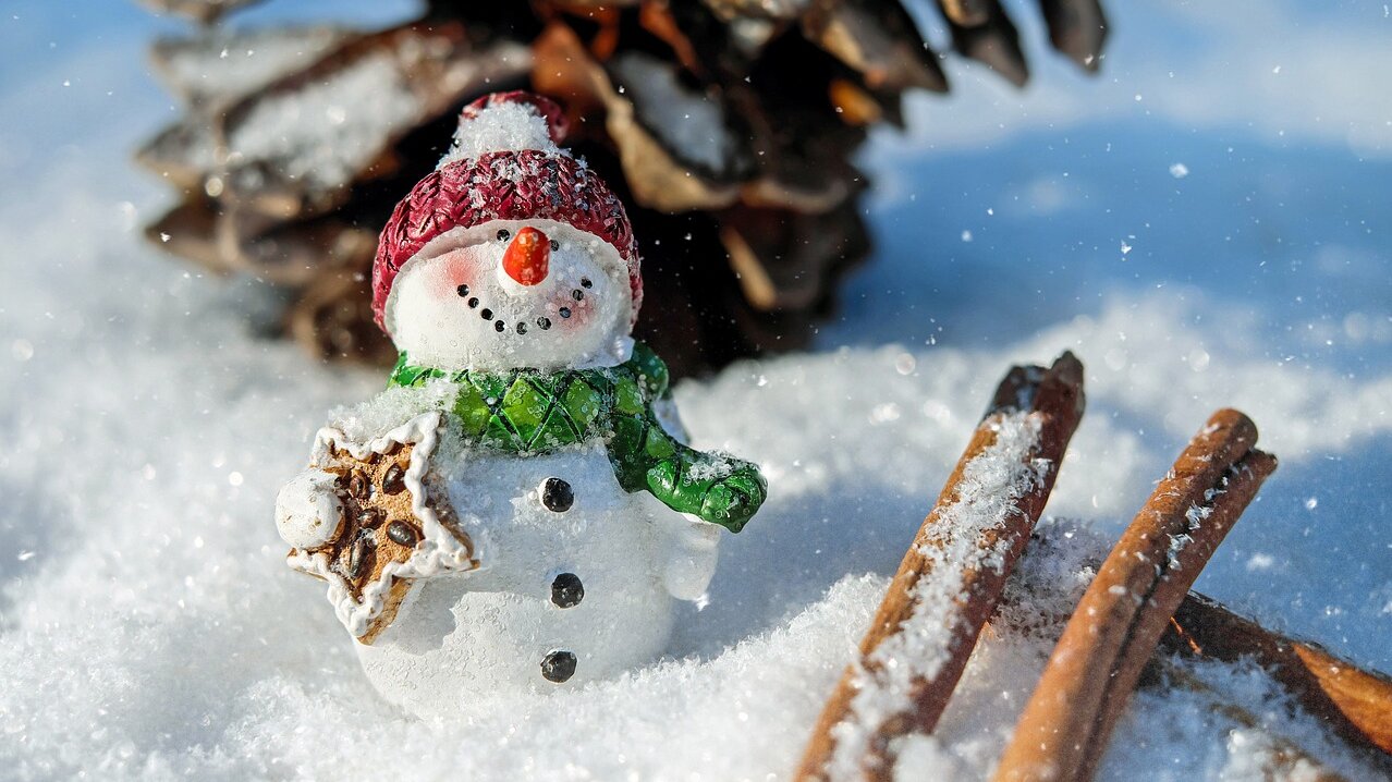 Ein kleiner Schneemann im Schnee. Daneben ein Tannzapfen und ein paar Zimtstangen.