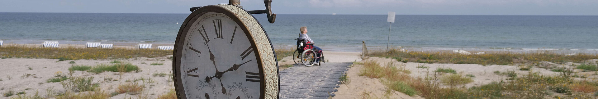 Ein Weg führt durch die Dünen zum Strand. Vorn am Weg eine Uhr. Am Ende des Weges, nahe am Wasser, eine Person im Rollstuhl.