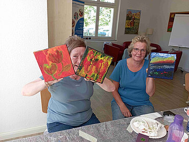 Zwei Frauen an einem Tisch mit Malutensilien halten Bilder in die Kamera.